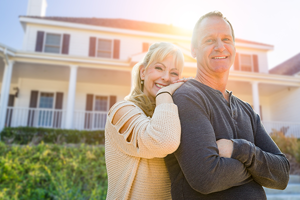 middle-aged Caucasian couple standing in front of their large suburban home smiling at the camera